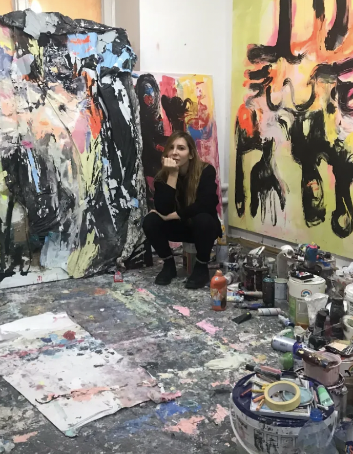 A person sits contemplatively in a vibrant, paint-splattered studio surrounded by abstract, colourful paintings, conveying a creative and chaotic atmosphere.