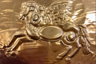 Golden embossed relief of a winged horse with intricate patterns. The metallic surface shines, giving a sense of majesty and craftsmanship.