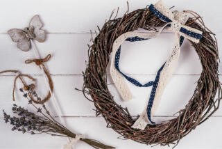 A rustic twig wreath adorned with lace and navy ribbon sits on a white wooden surface. Nearby are a butterfly decoration and a bundle of lavender, creating an elegant, handcrafted feel.