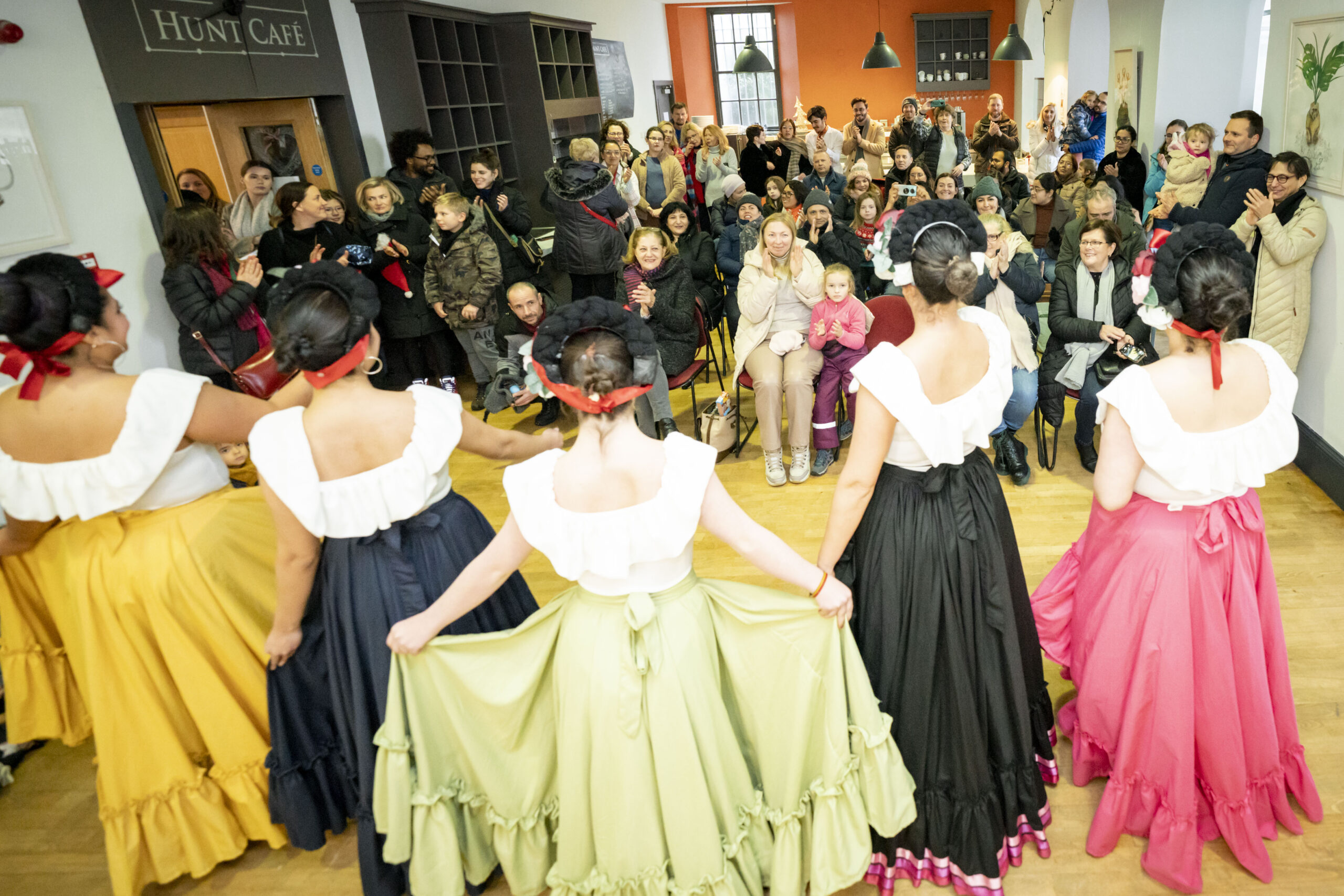 Five dancers in colourful skirts and white tops perform in front of a smiling audience in a lively café setting. The room is filled with warmth and applause.