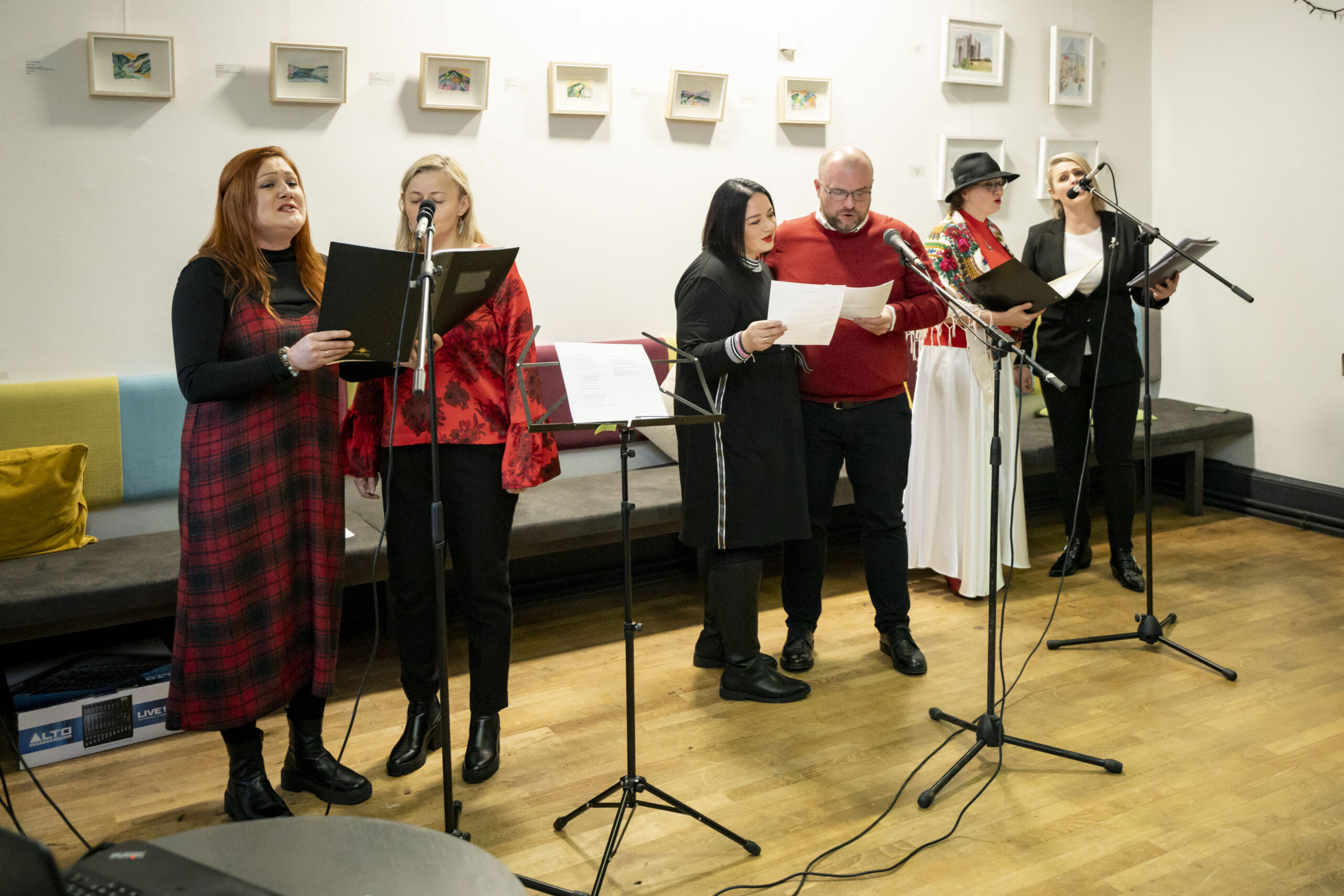A diverse group of six people sings together in a cosy room, holding sheets and standing at microphones. The scene feels lively and collaborative, with bright art on the walls.