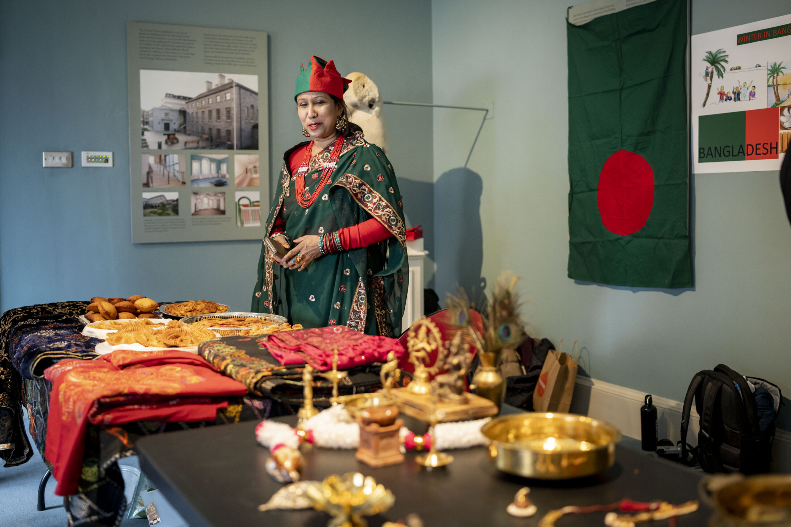 A woman in traditional Bangladeshi attire stands by a table with various foods and ornate items. A Bangladesh flag hangs on the wall, conveying cultural pride.