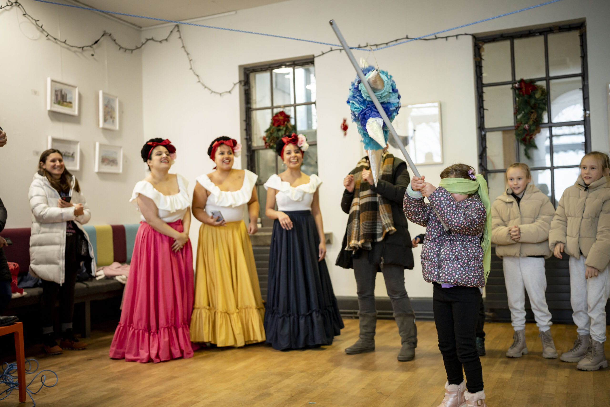 A blindfolded child swings at a colourful piñata in a festive room. Three women in vibrant traditional dresses watch and smile, while others stand nearby, creating a joyful atmosphere.