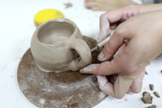 Hands shaping a clay mug on a pottery wheel. The scene conveys focus and creativity, with clay pieces and a sponge in the background.