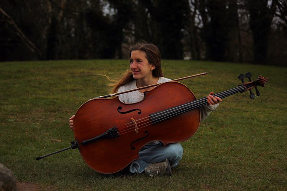 A young person sits on grass, smiling while holding a large cello horizontally. The setting is outdoors with trees in the background, creating a serene and joyful atmosphere.