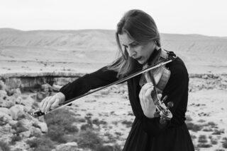 A woman in a black dress passionately plays the violin in a vast, open desert landscape, conveying a sense of serenity and focus.