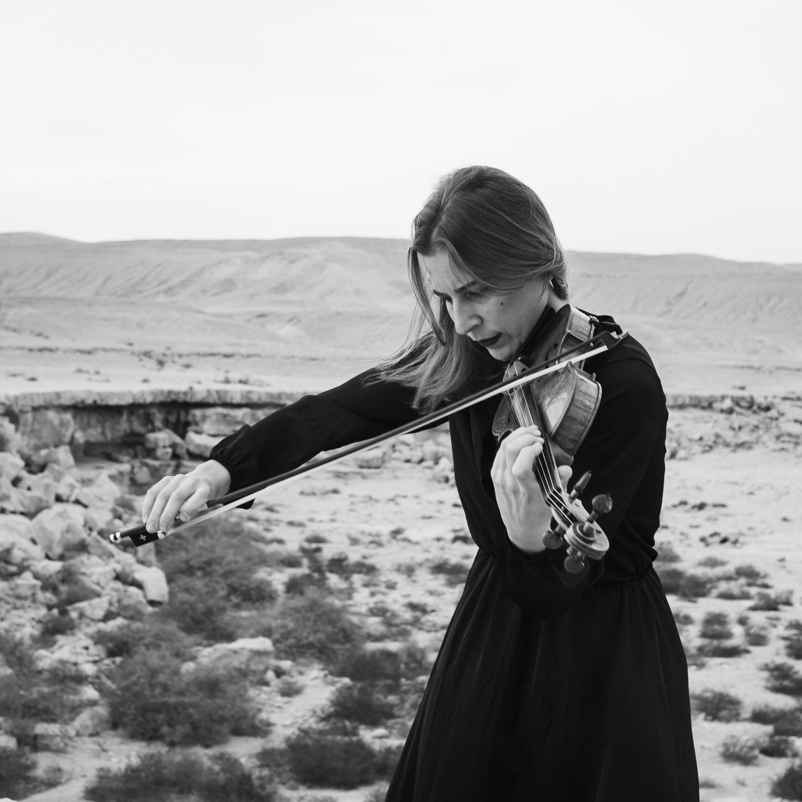 A woman in a black dress passionately plays the violin in a vast, open desert landscape, conveying a sense of serenity and focus.