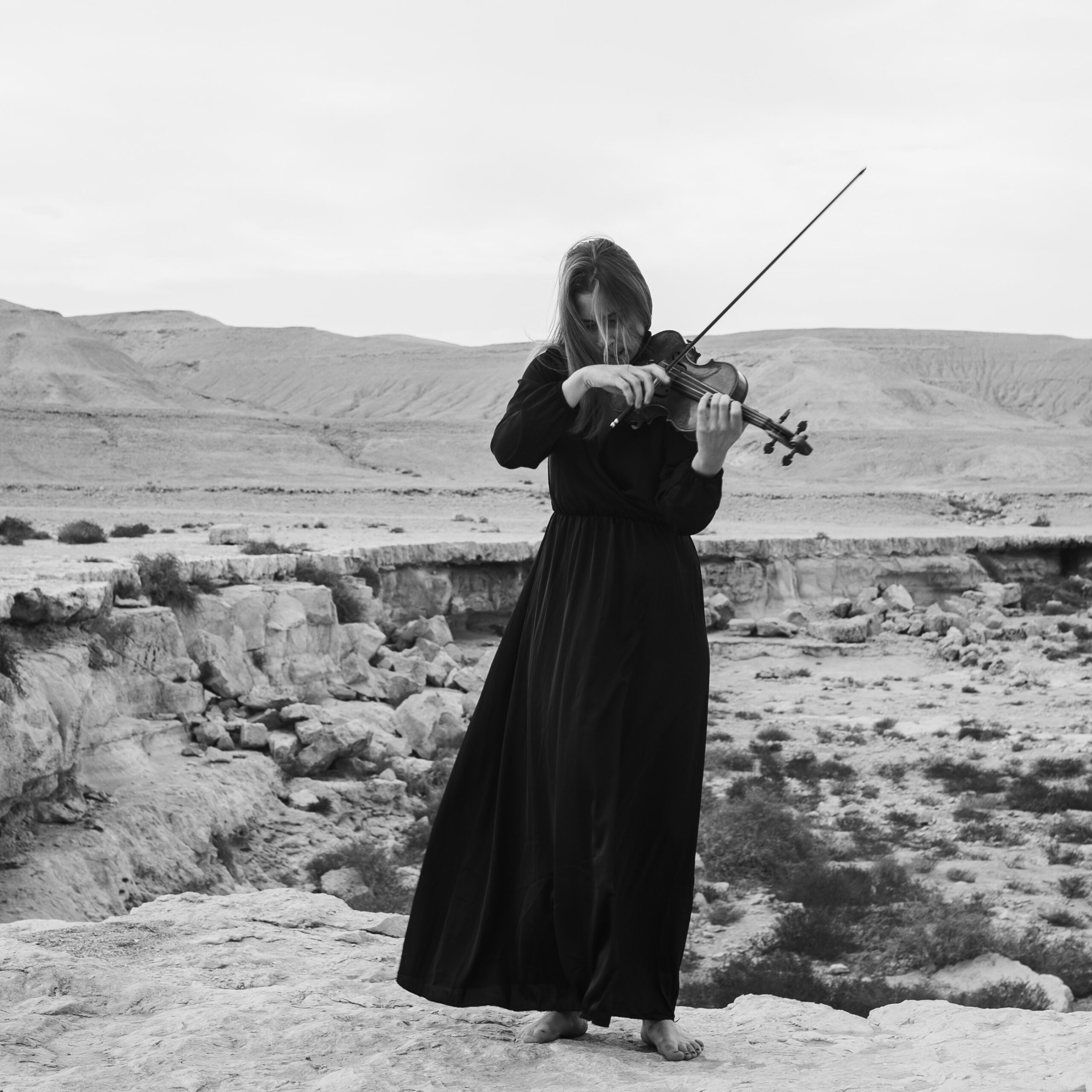 A woman in a long black dress plays the violin on a rocky desert landscape. The black-and-white image conveys a serene, introspective mood.