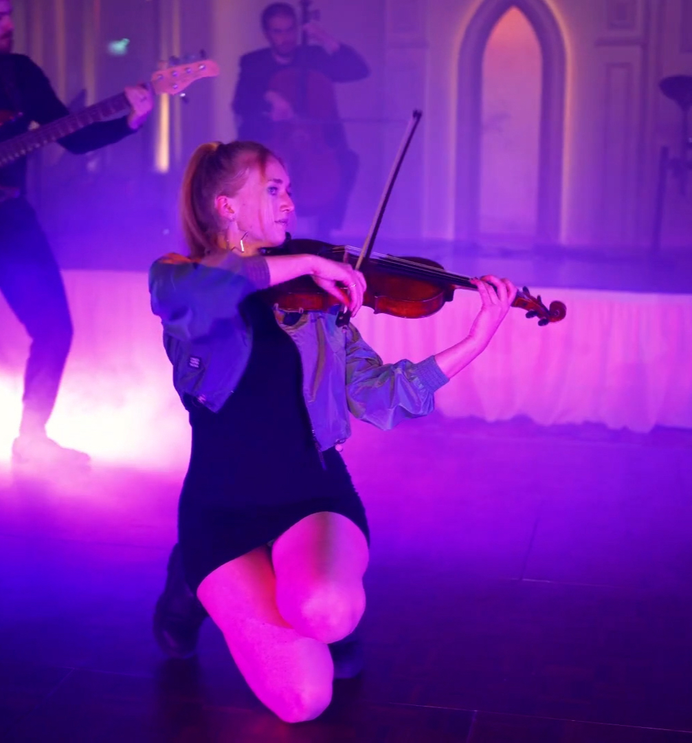 A woman kneels, playing the violin passionately on a misty stage with vibrant purple lighting. Two musicians in the background add depth to the energetic scene.