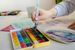 A hand dipping a paintbrush into water colours