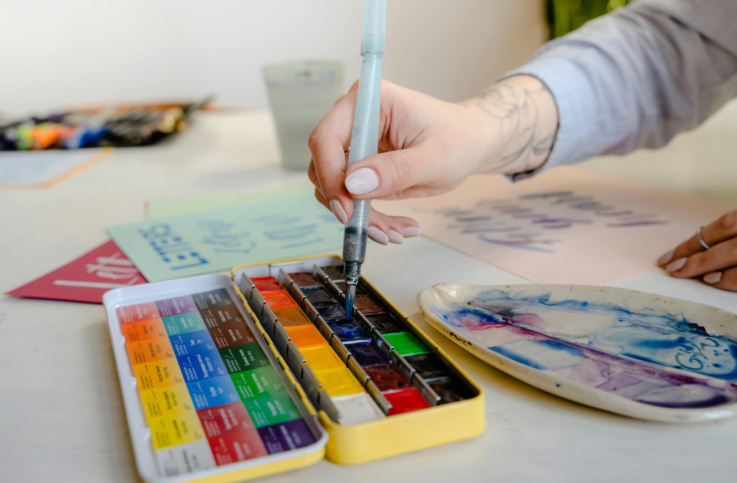 A hand dipping a paintbrush into water colours