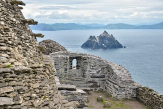 Ancient stone ruins overlook the ocean, with a rugged island emerging from the water against a backdrop of distant green hills.
