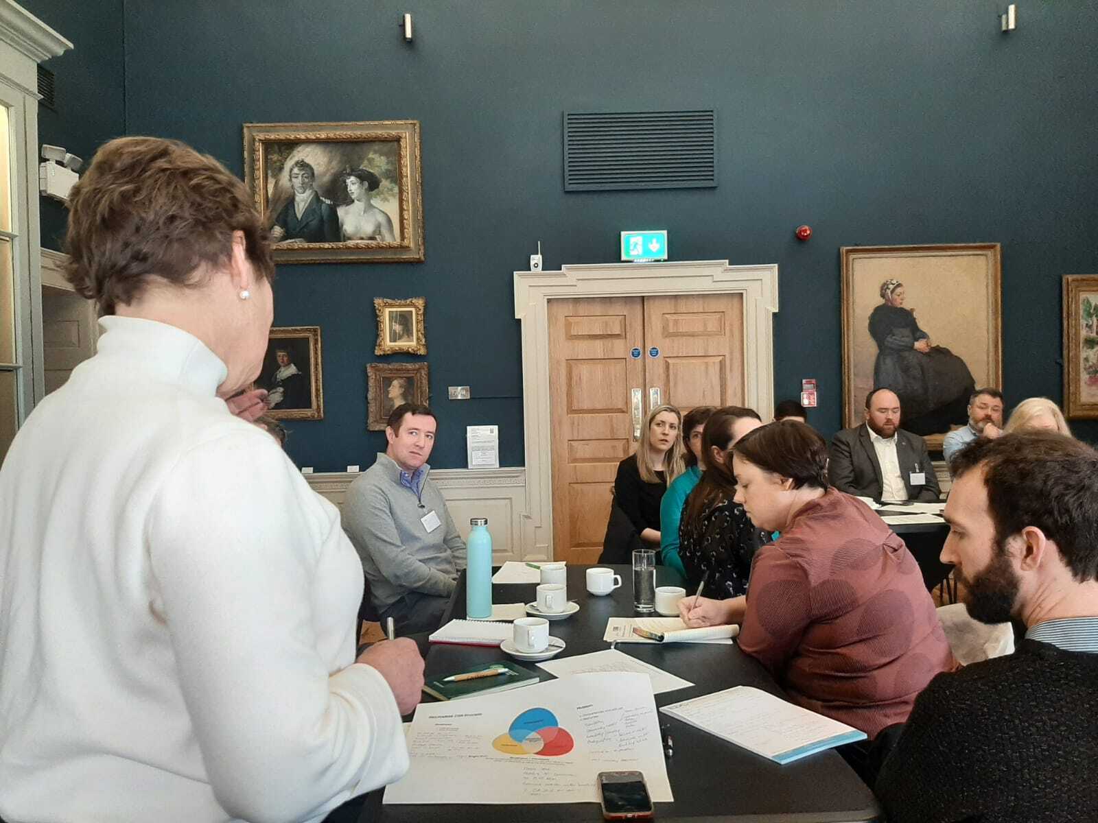 A professional meeting in a gallery, with participants engaged in discussion around a table covered in papers and cups.