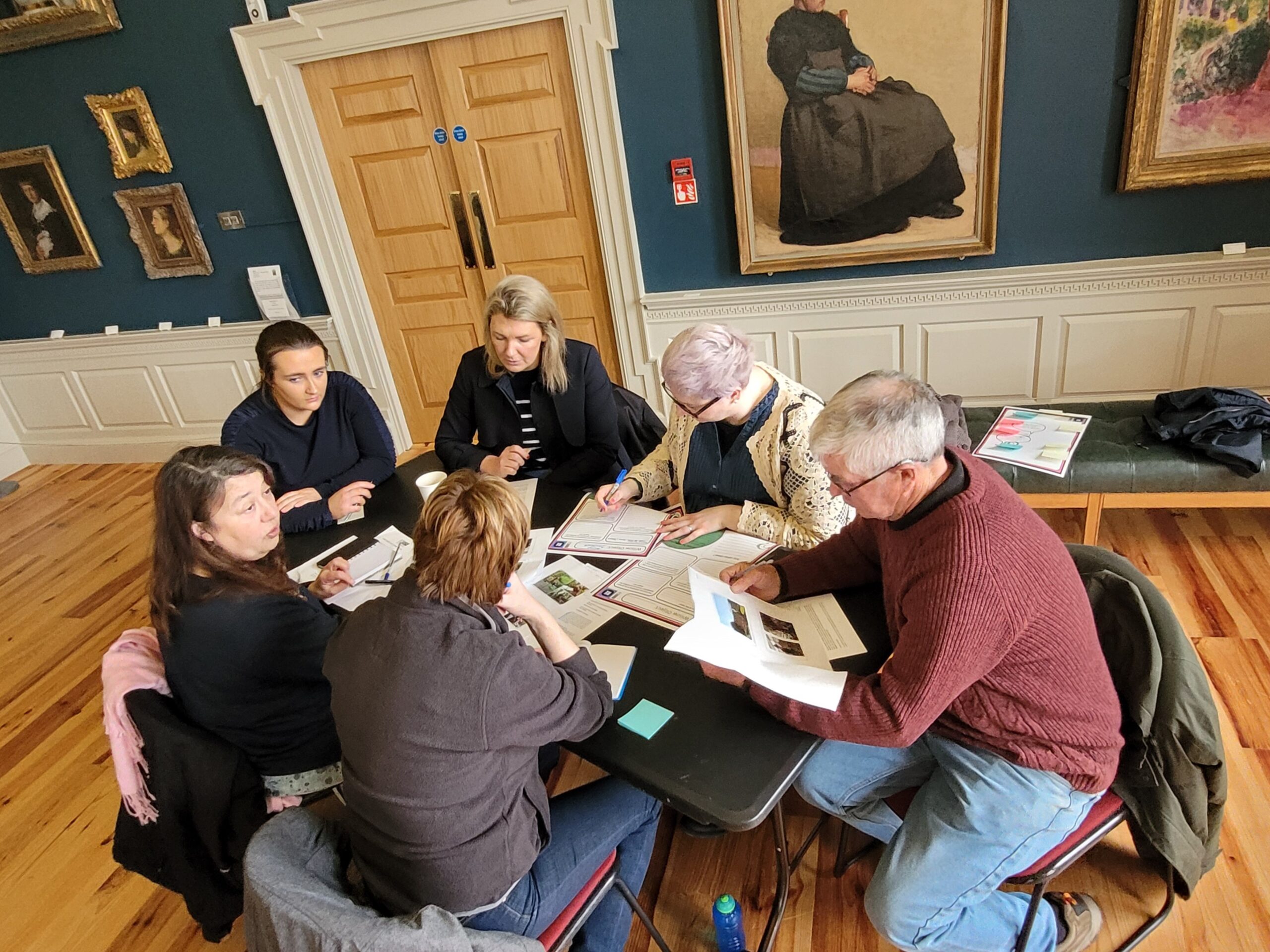 A group of six people gathered around a table, discussing and reviewing documents in a warmly lit art gallery.