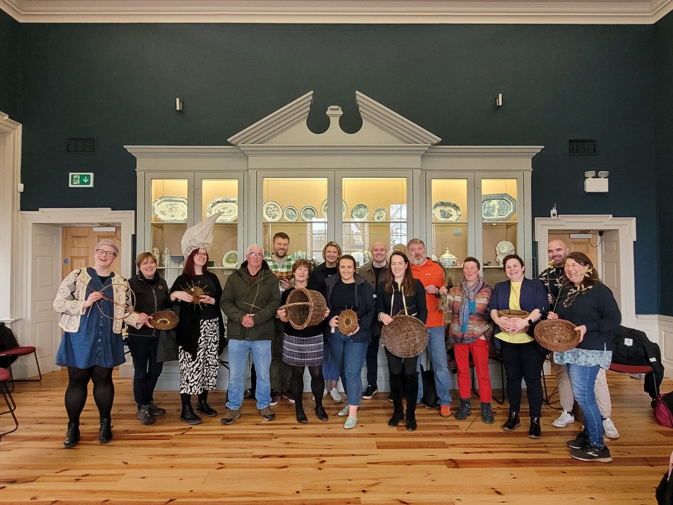 A diverse group of people stands in a museum, proudly holding handmade baskets, with decorative ceramics displayed behind them.