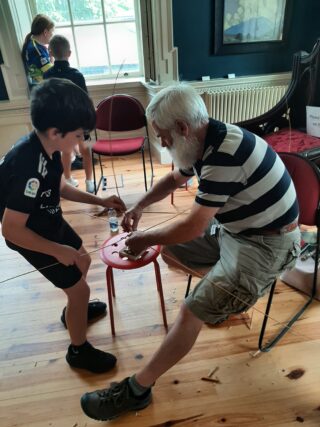 An elderly man and a child collaborate on a craft project using sticks and materials on a small chair in a well-lit room.