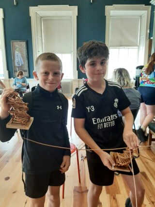 Two boys hold crafted items made of twigs, smiling in a bright, colourful room filled with other children engaged in activities.