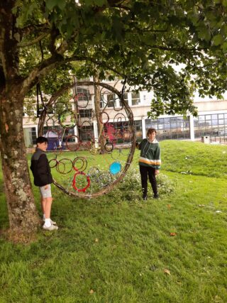 Two people stand near a large, colourful circular structure made of intertwined ropes, set against a backdrop of grass and trees.