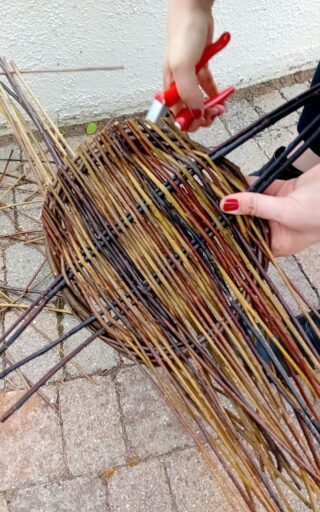 A person's hand with red nail polish holds scissors, trimming colourful woven branches into a decorative basket shape on stone pavement.