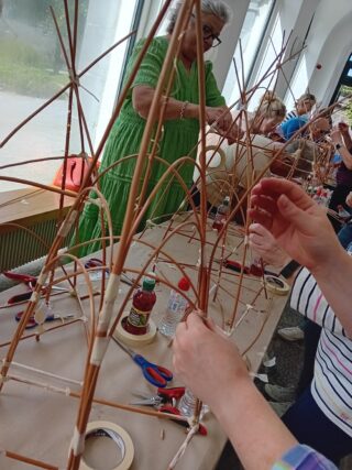 A group of people engages in a crafting workshop, creating structures from natural materials at a long table filled with supplies.