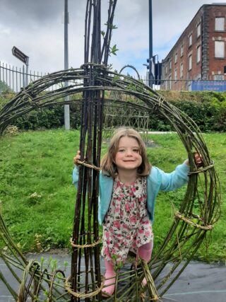 A child in a floral dress and blue cardigan plays in a grassy area, interacting with a large, circular woven structure.