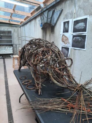 A handwoven animal sculpture made of twigs is displayed on a table in a studio with shading from skylights and artwork on the wall.