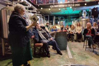A speaker holds a book in a brewery while an audience listens intently, surrounded by large metal brewing tanks and ambient lighting.
