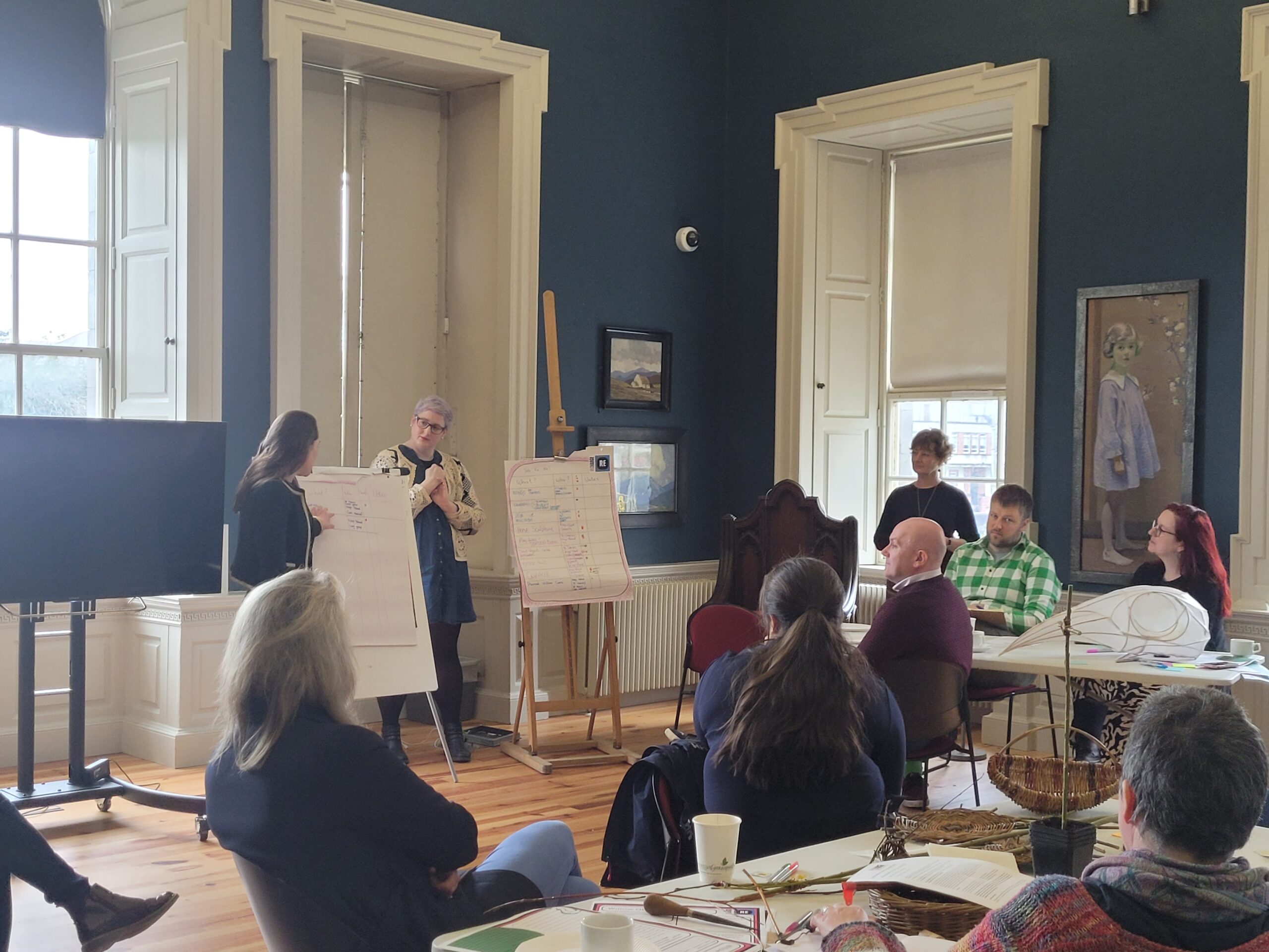 A group participates in a workshop, observing a presenter at a flip chart in a sunlit room with blue walls and large windows.