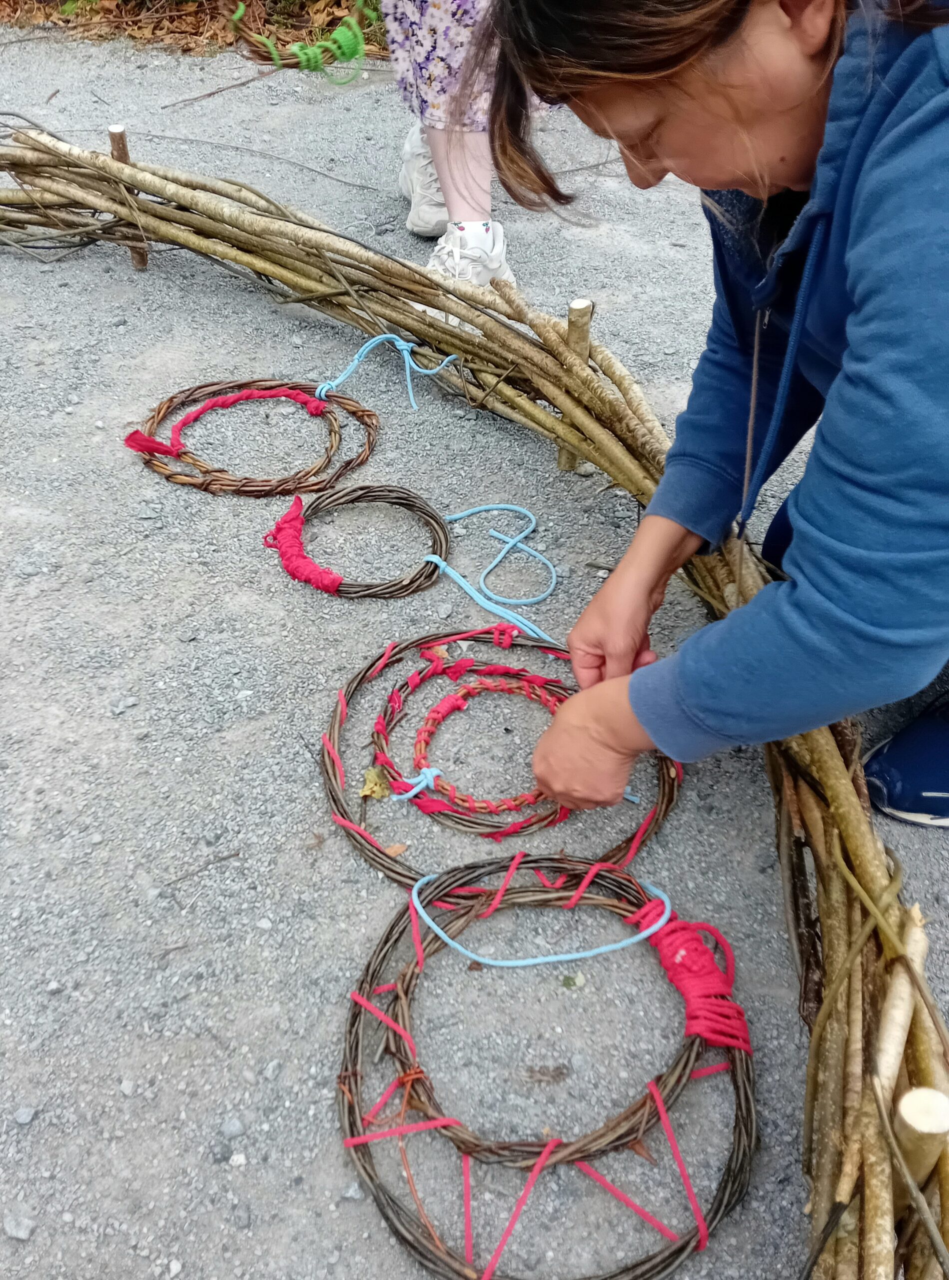 A person creates woven circular decorations from twigs and colourful string, surrounded by additional materials on a gravel surface.