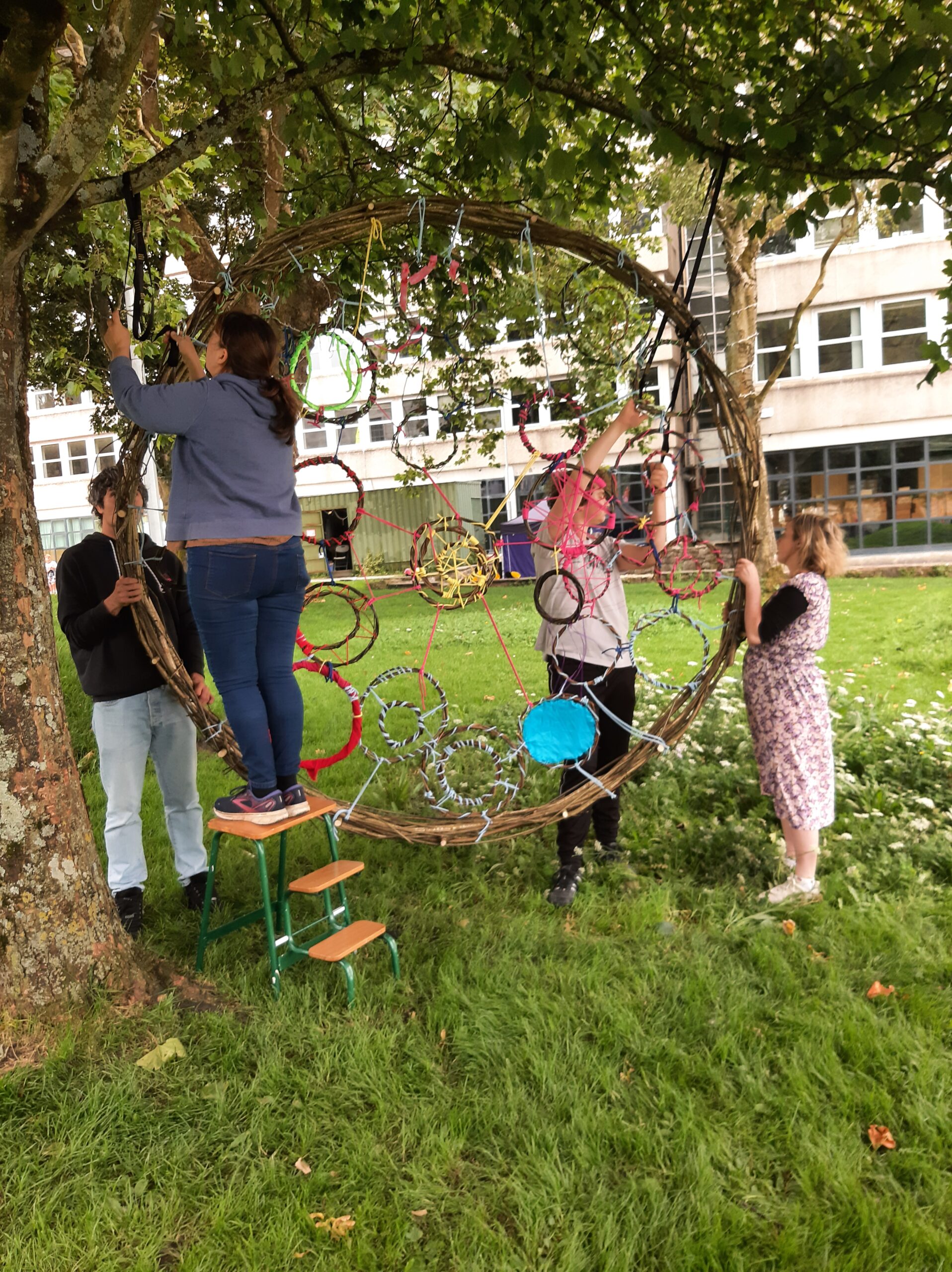 A group of four people works together to hang a large, colourful circular art piece from a tree in a grassy park setting.