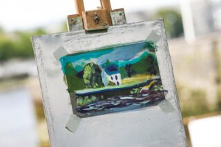 A colourful landscape painting of a house by a river, displayed on an easel with masking tape at the corners, outdoors.