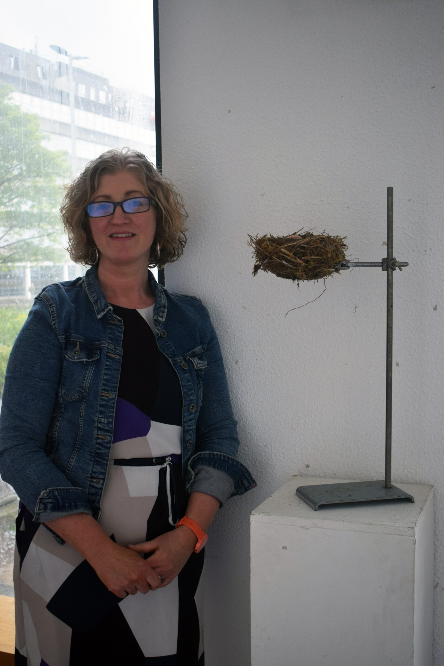 A woman stands beside a unique art installation featuring a small bird's nest mounted on a metal stand against a light wall.