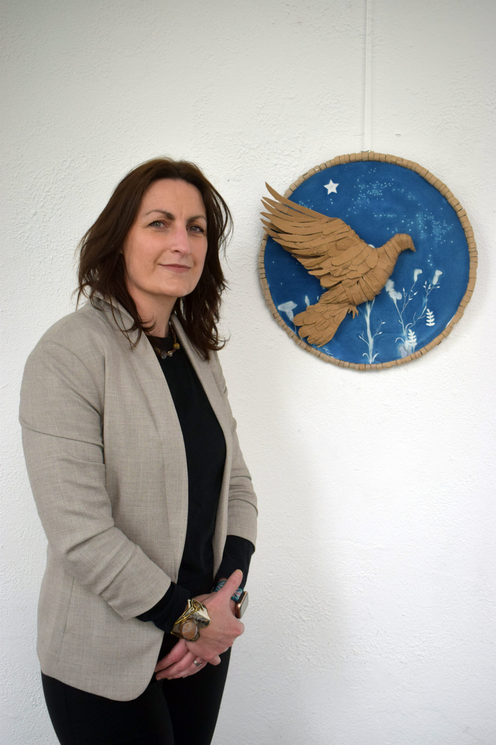 A woman in a light blazer stands next to a circular blue wall art featuring a gold bird in flight against a textured white background.