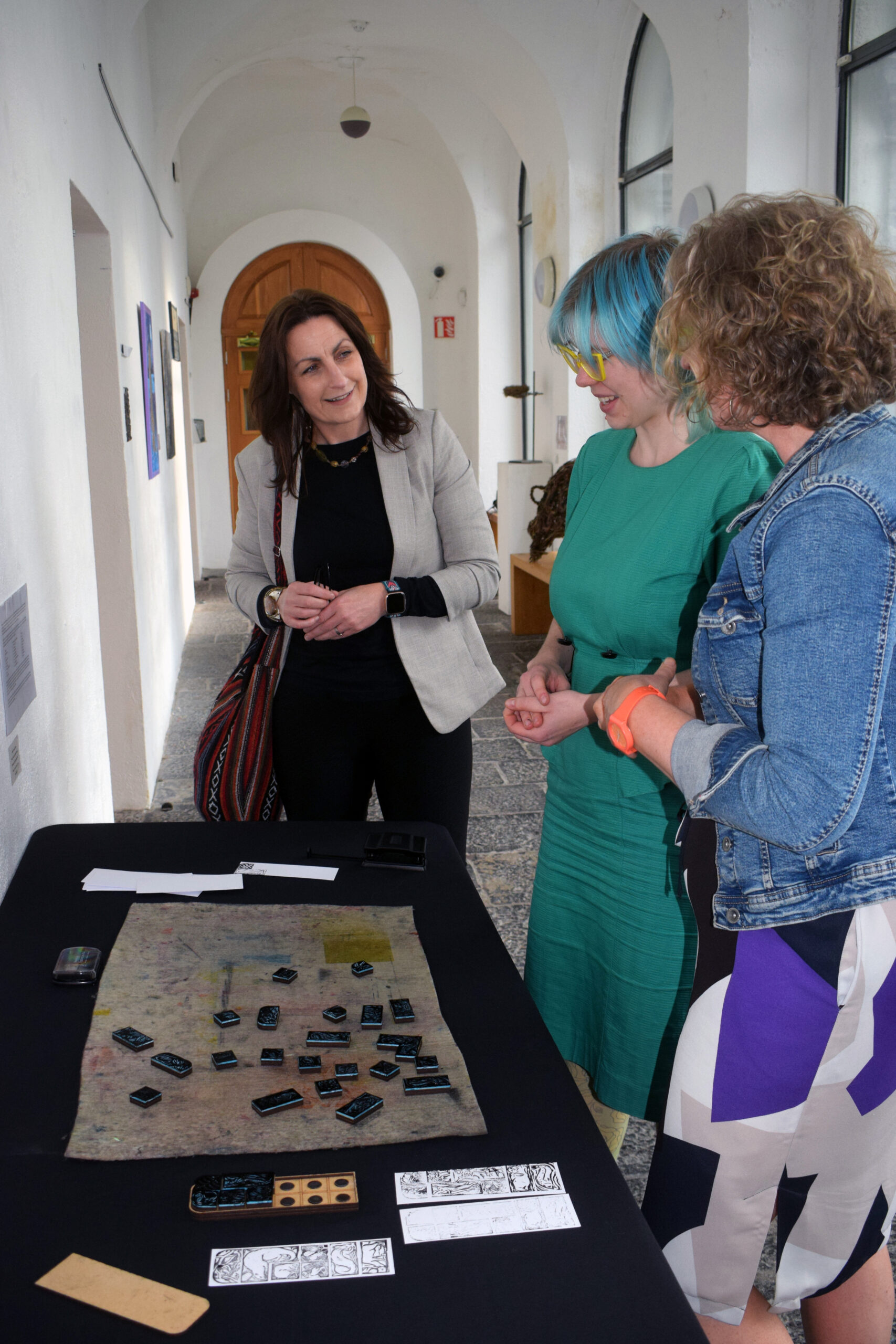 Three women engage in discussion around a table with art materials, including various stamps and printed designs, in a gallery hallway.