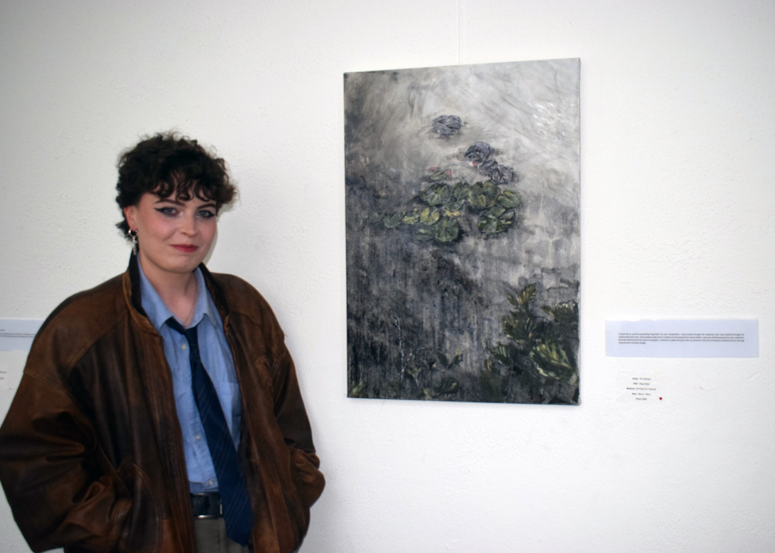 A person wearing a brown leather jacket stands beside a muted painting of lily pads on a grey background in an art gallery.