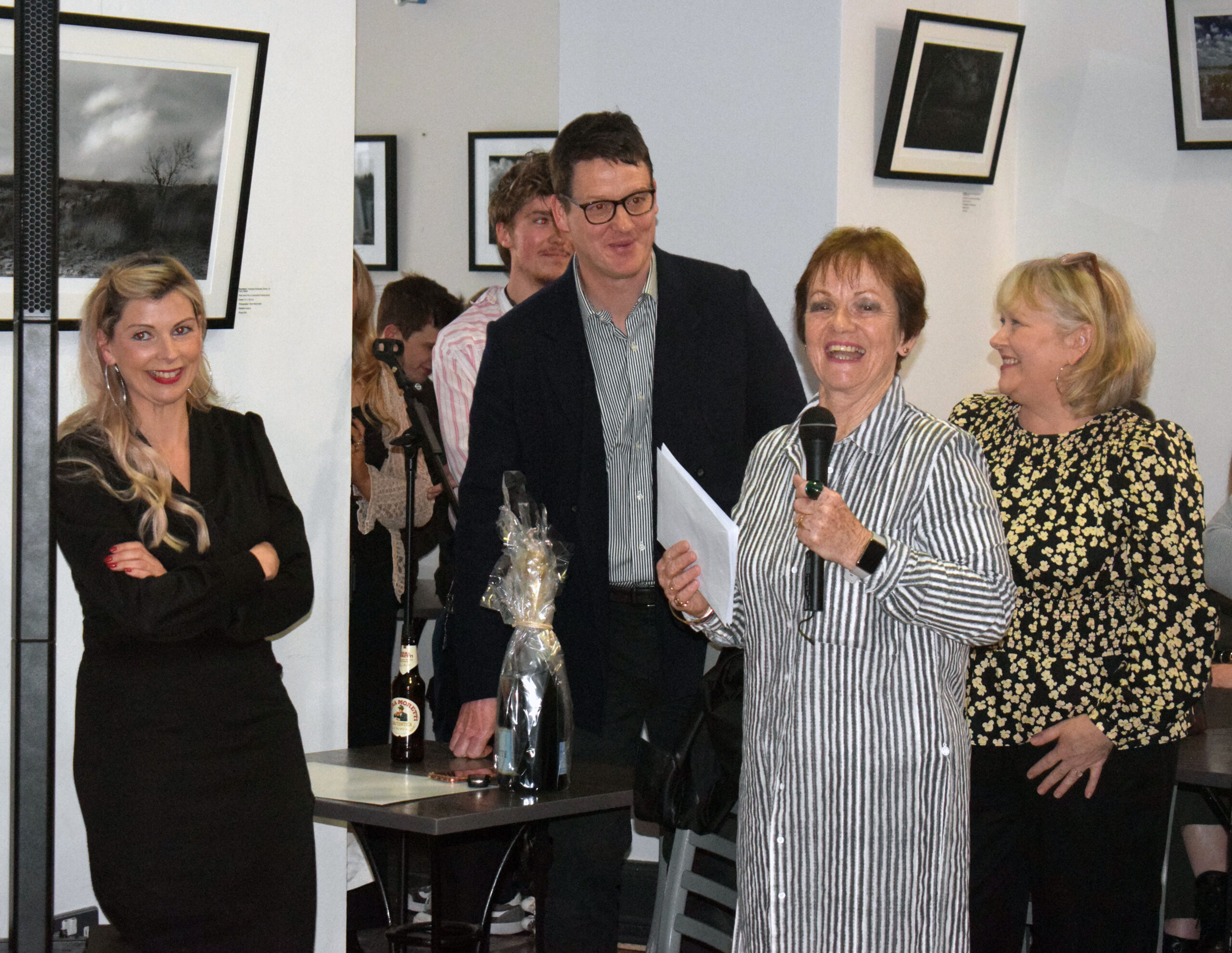 A woman holding a microphone speaks with another woman in a striped dress during an event surrounded by onlookers and artworks.