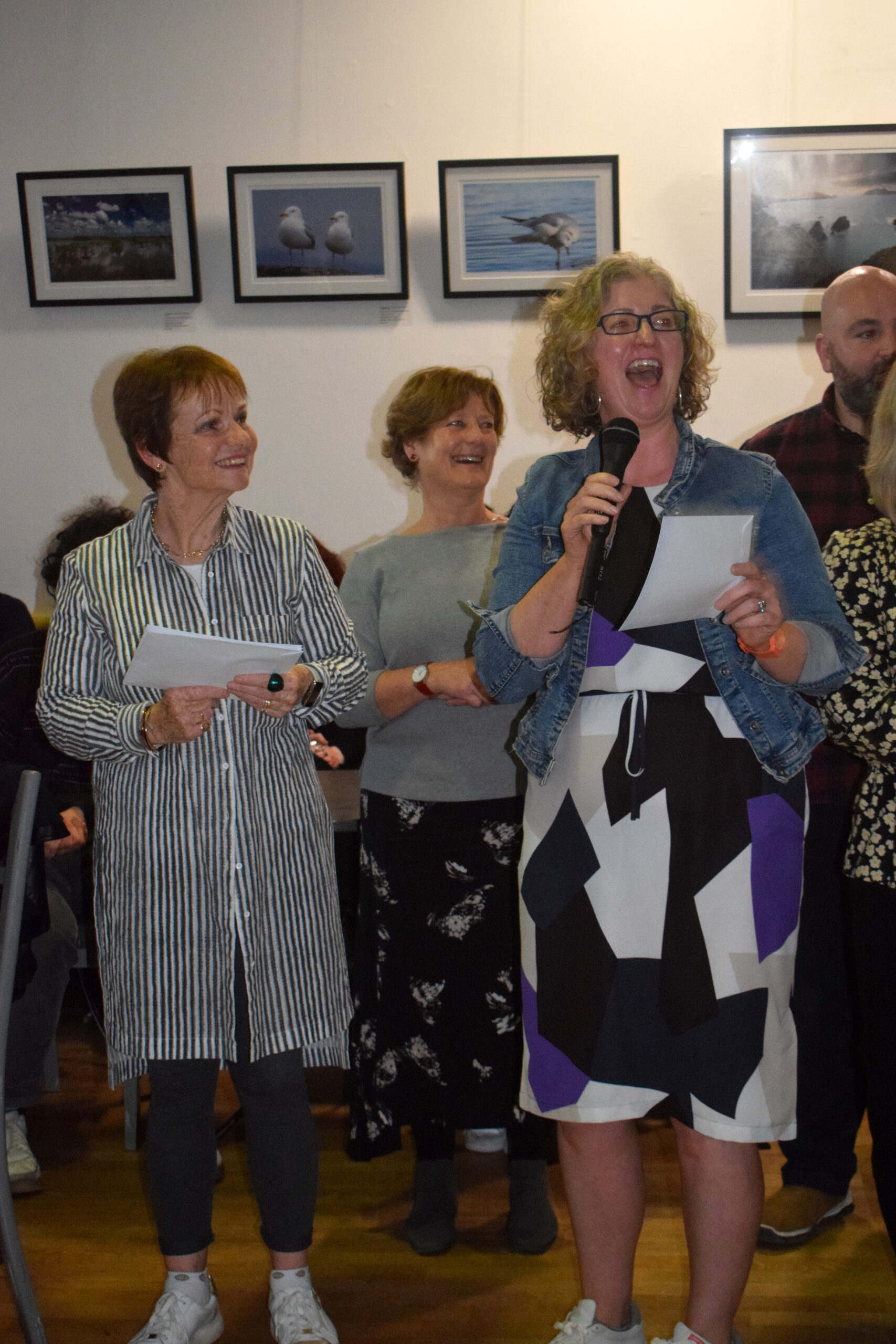A group of women stands together, holding papers, while one speaks into a microphone. Framed nature photographs adorn the wall behind them.
