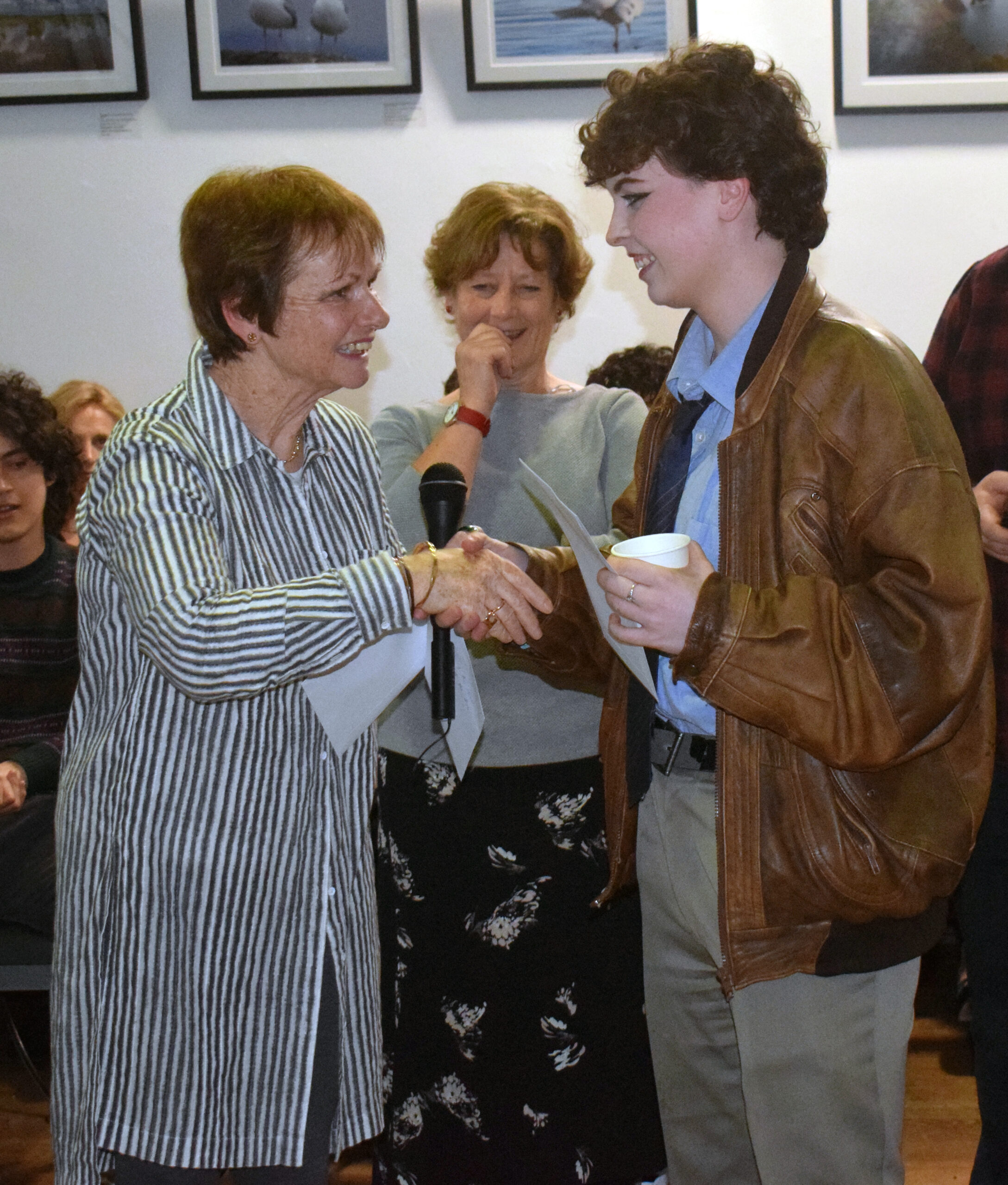 Two individuals shake hands at an event, surrounded by a small crowd and photography displayed on the walls. One holds a cup.