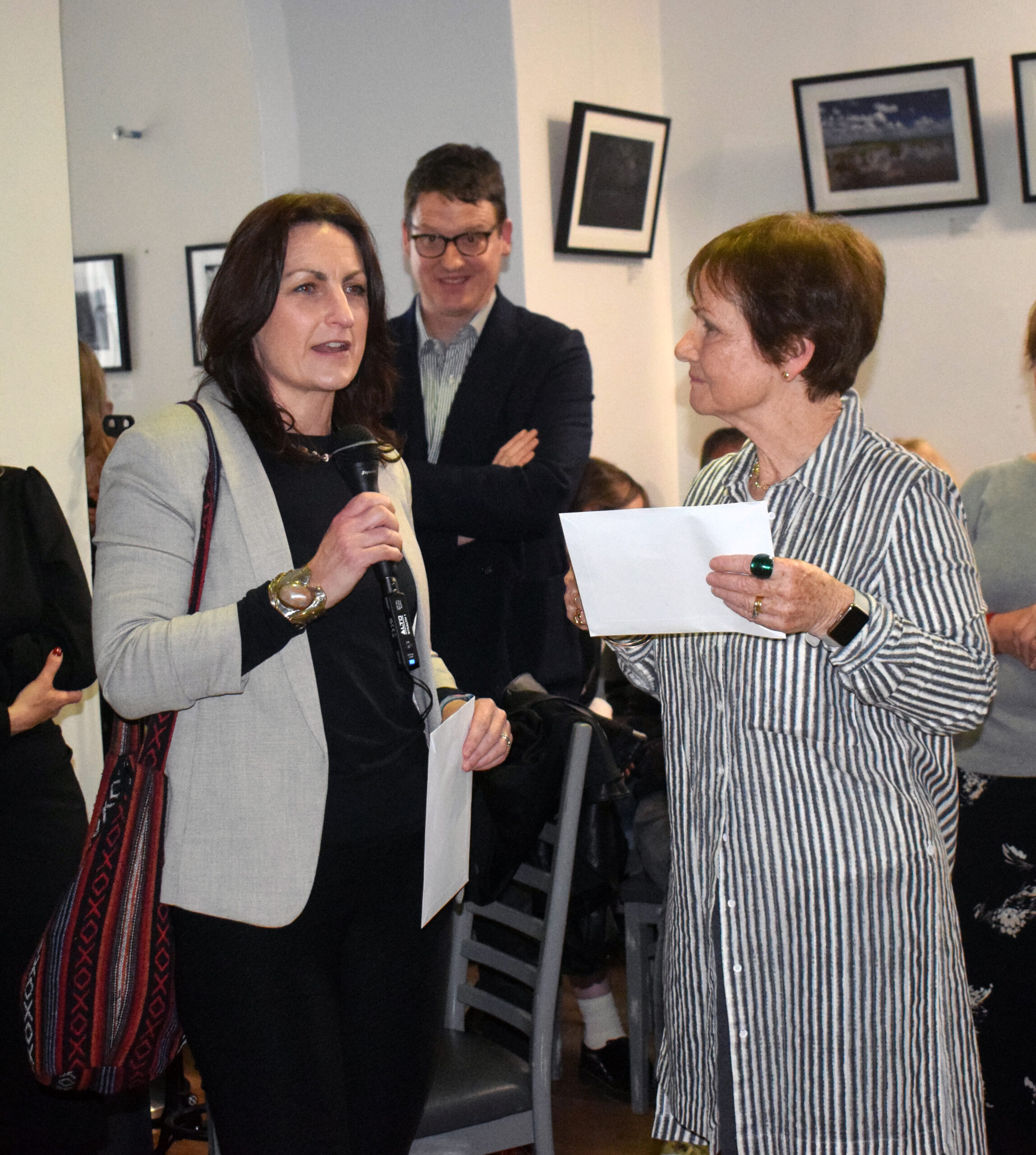 A woman stands with a microphone, engaging in conversation with another woman holding a document, surrounded by an attentive audience in a gallery.