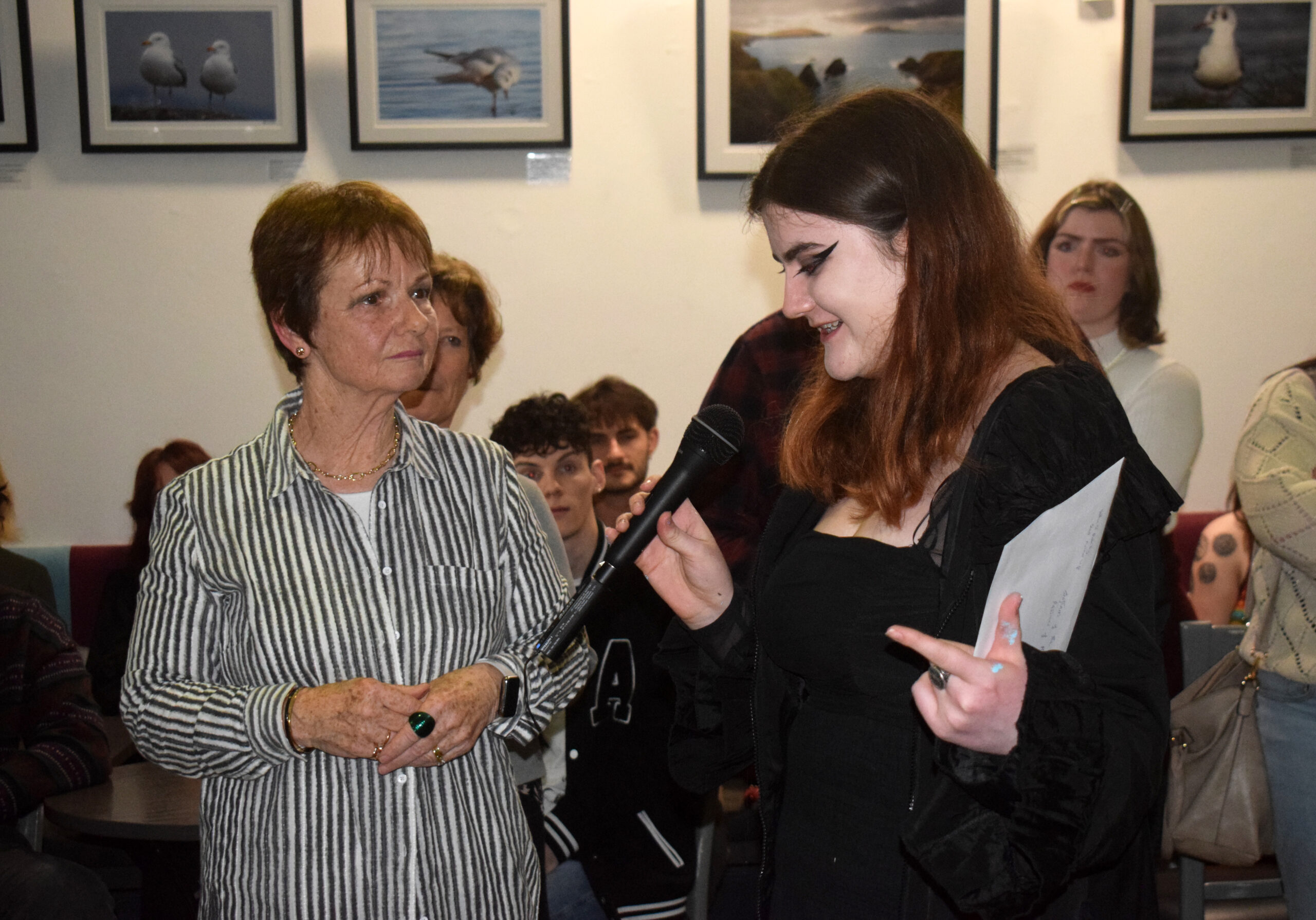 A woman holds a microphone and gestures while speaking to another woman in a casual gathering surrounded by framed bird photographs.