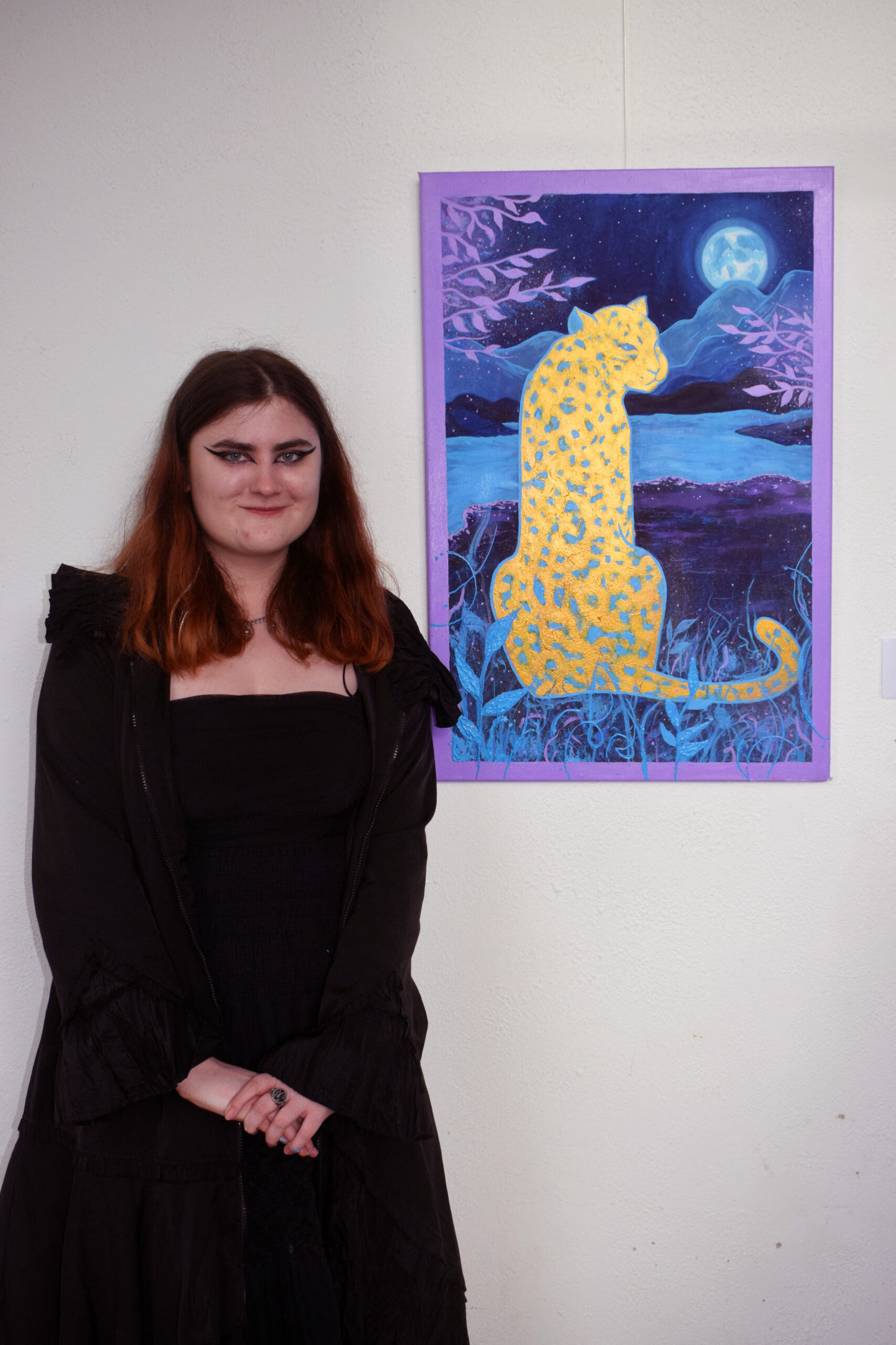 A person in a black outfit stands beside a vibrant artwork of a golden leopard against a night-time landscape background.