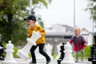 Two children play with oversized chess pieces in a park, with greenery and buildings blurred in the background.