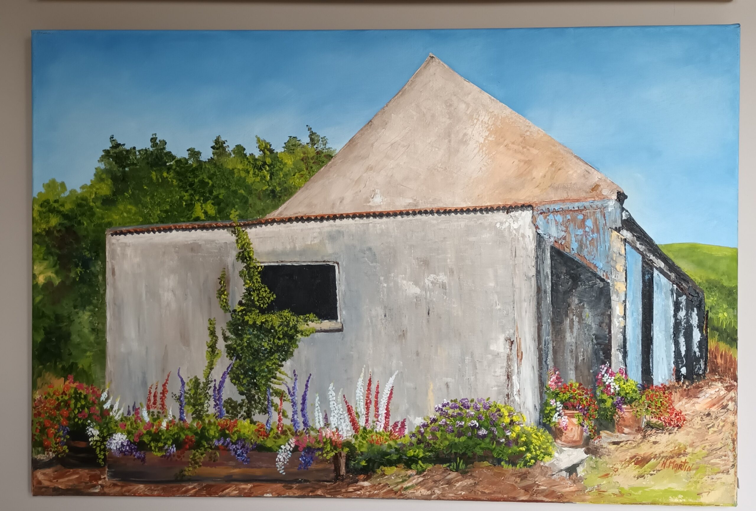 A weathered farmhouse with a sloped roof, surrounded by vibrant flowers and greenery under a clear blue sky.