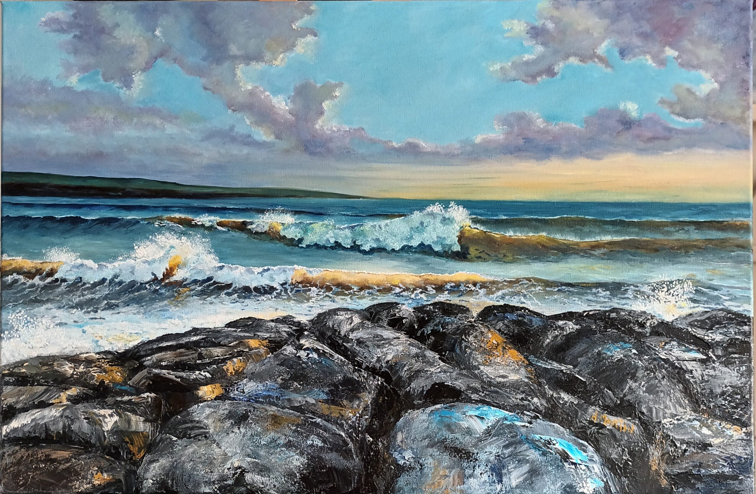 A scenic ocean view with rolling waves crashing against dark rocks under a bright blue sky, accented by fluffy clouds at sunset.