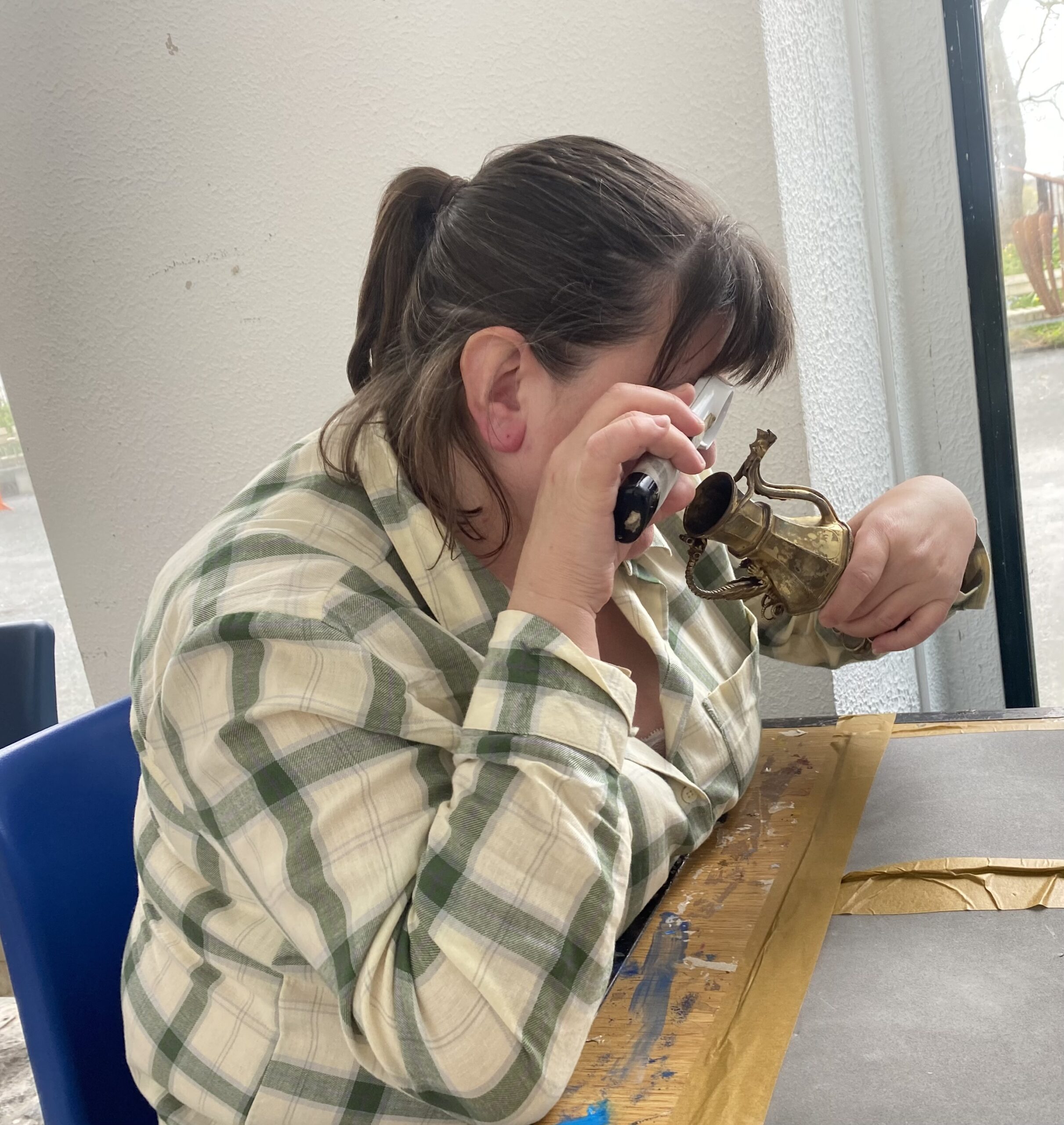 A person with long hair, wearing a green and white checked shirt, holds a brass tool while seated at a table with art supplies.
