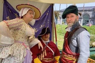 Three individuals in historical costumes pose in a tent, showcasing attire from a bygone era, with a green cannon in the background.