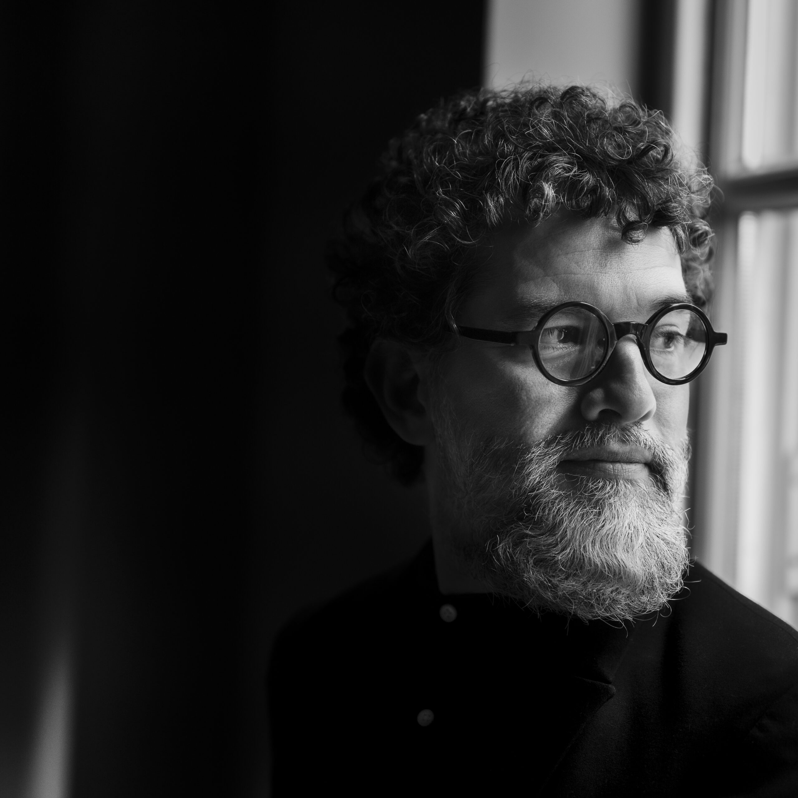 A person with curly hair wearing a dark shirt stands against a softly lit background, capturing a moment in monochrome.