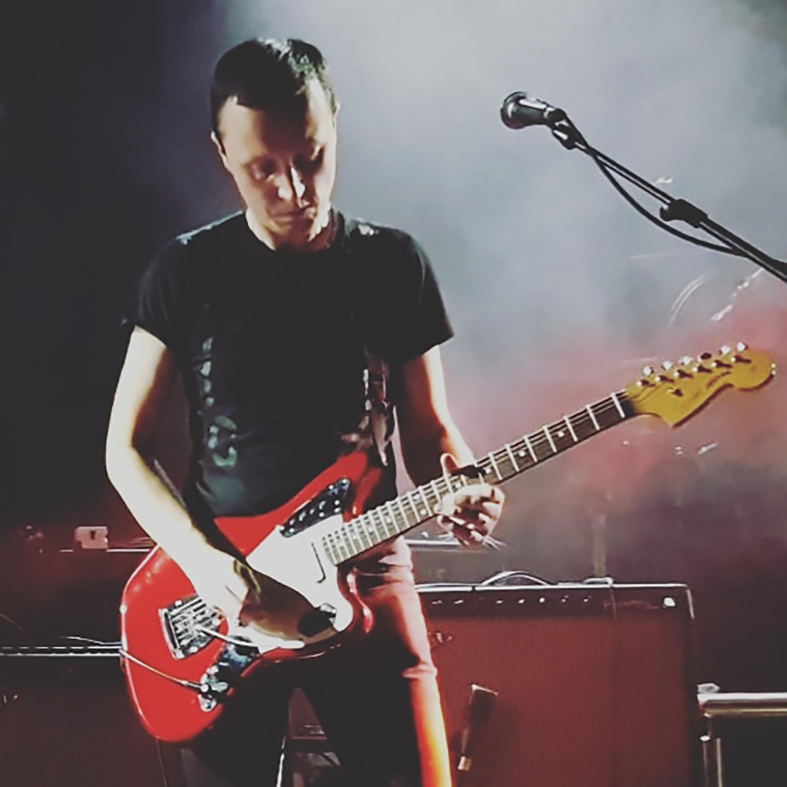 A musician plays an electric guitar on stage, surrounded by atmospheric lighting and equipment, creating an engaging live performance scene.