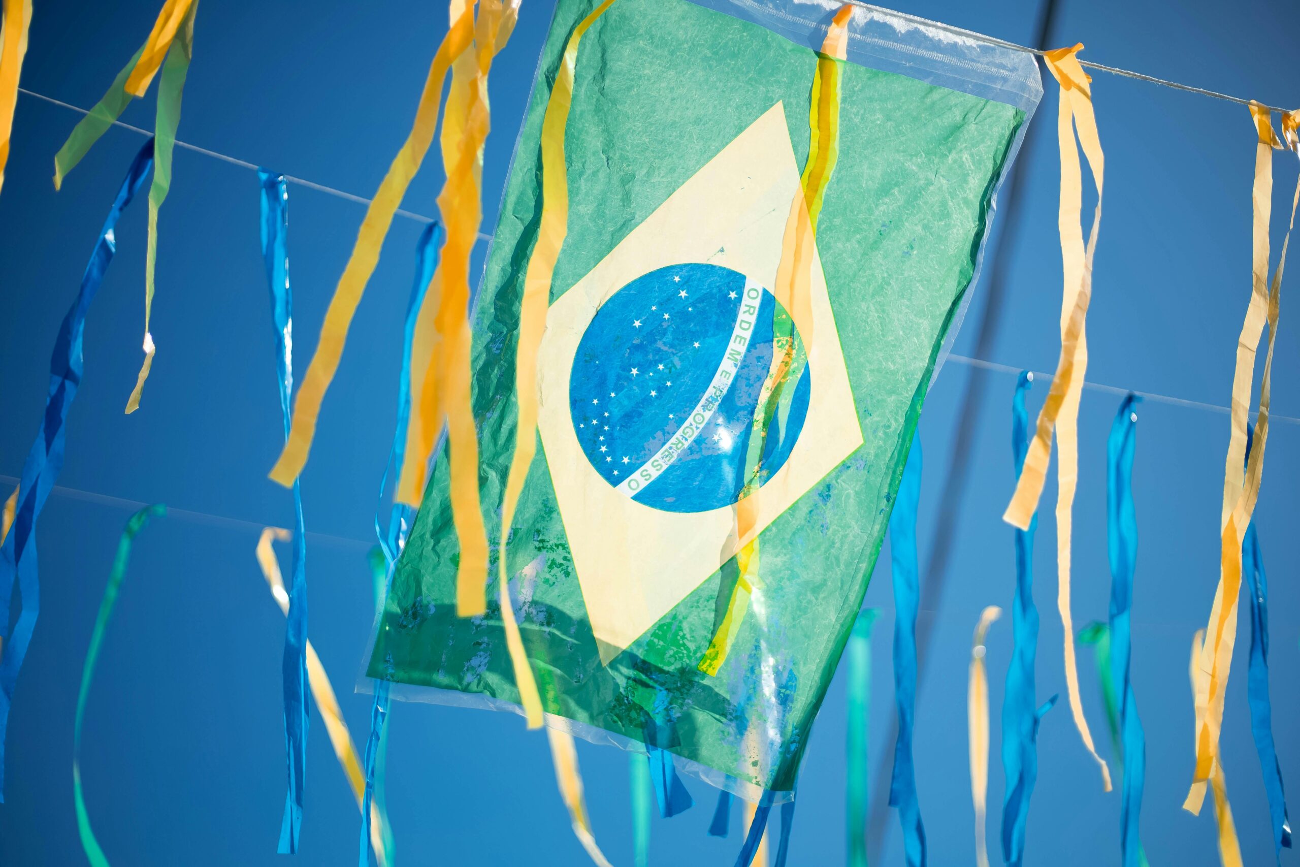 A Brazilian flag hangs between colourful blue and yellow streamers against a bright blue sky.