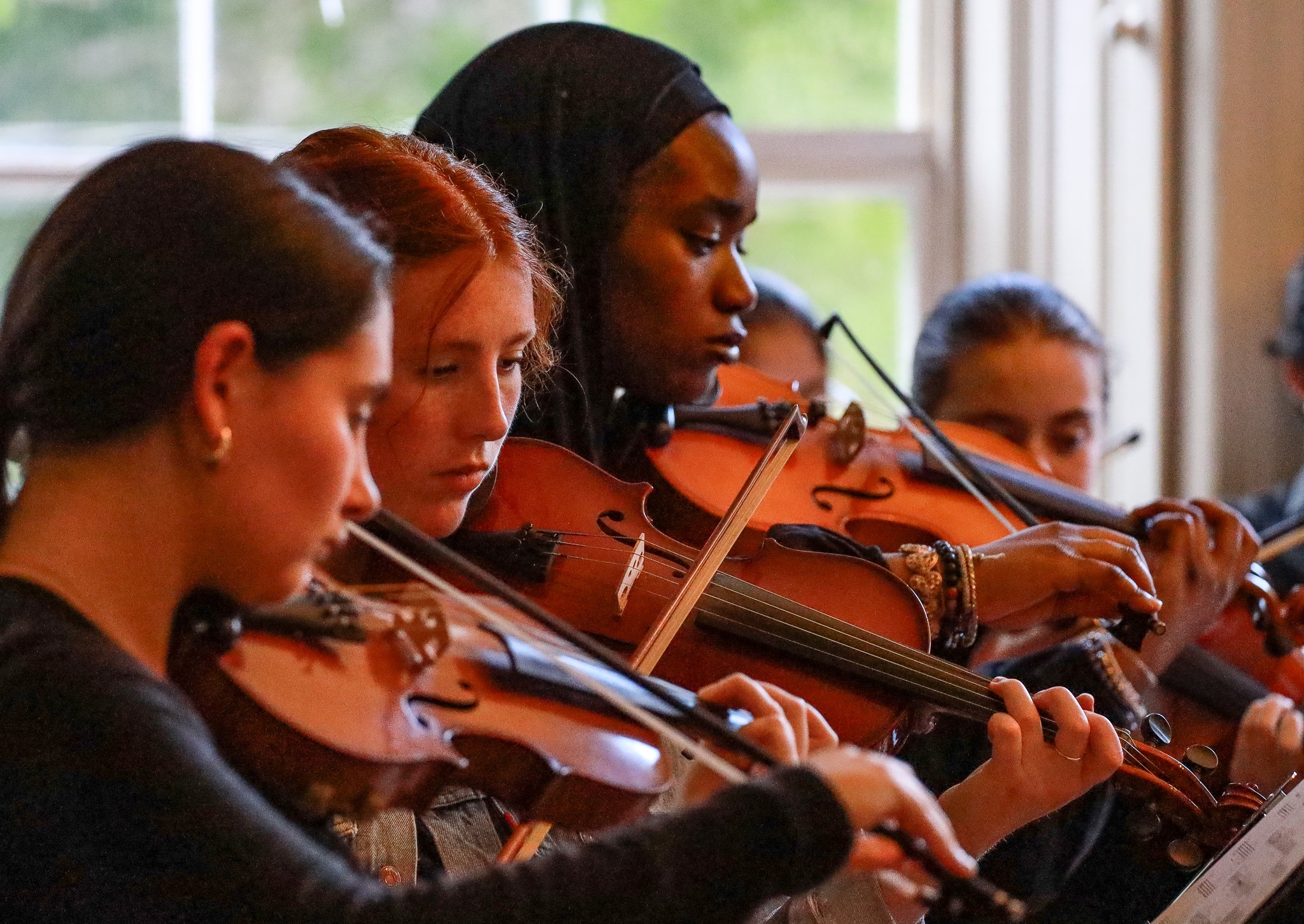 A group of musicians playing violins, focused on their instruments in a bright room with large windows.